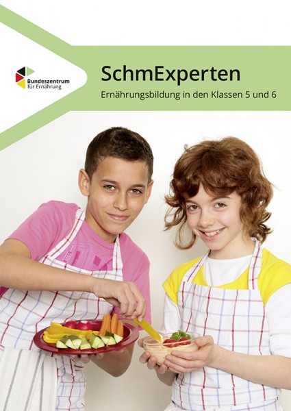 Zwei Kinder, die Schürzen tragen, halten einen Teller mit Gemüse-Sticks und Hummus und lächeln