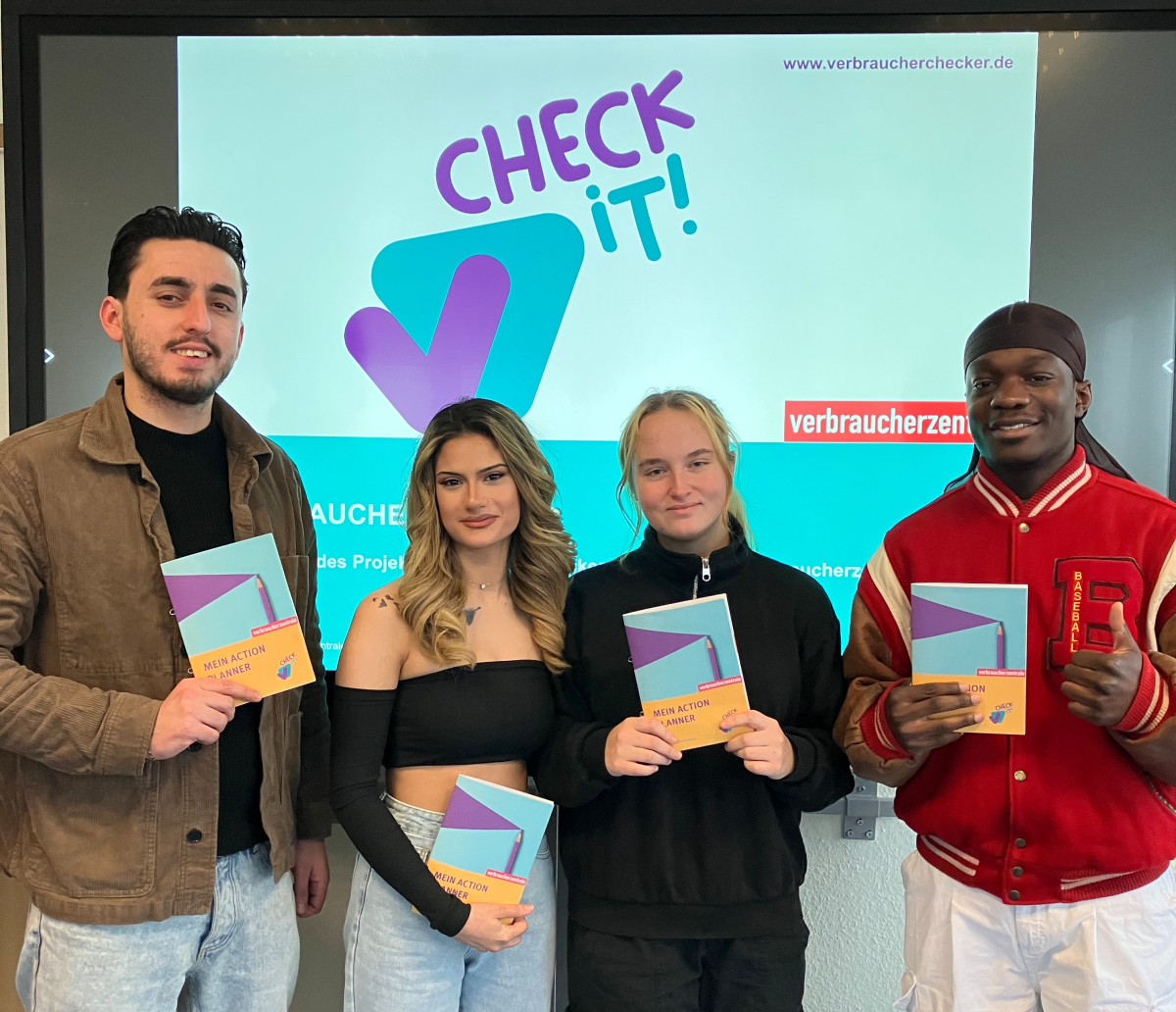 Vier junge Erwachsene stehen vor einem Smartboard, auf dem das Check-it-Visual der Verbraucherchecker und das Logo der Verbraucherzentrale zu sehen sind. Sie halten den Action Planner, das Lernmaterial der Peer-Scout-Ausbildung, in ihren Händen und lächeln in die Kamera.