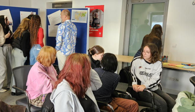 Junge Personen informieren sich an einer Stellwand mit Informationen, andere diskutieren in einer Gruppe.