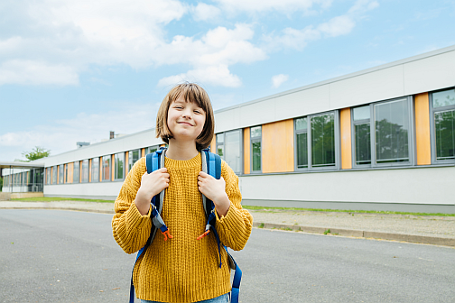 Ein Kind im gelben Pullover steht bei gutem Wetter stolz und fröhlich vor einem Schulgebäude.
