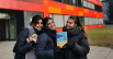 Drei junge Frauen stehen vor einem Schulgebäude, halten den Action Planner hoch und lächeln in die Kamera.