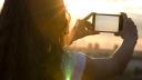 Eine Person mit langen Haaren hält ihr Smartphone im Querformat vor sich und fotografiert den Himmel während eines Sonnenuntergangs