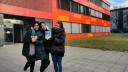 Drei junge Frauen stehen vor einem roten Gebäude und halten lächelnd ein Heft in die Kamera.