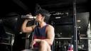 Ein junger muskulöser Mann in Sportkleidung sitzt in einem Fitnessstudio vor einem Gerät und trinkt aus einer Trinkflasche.
