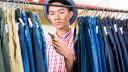 Ein junger Mann mit Hut steht in einem Bekleidungsgeschäft und schaut Hosen auf einer Kleiderstange an. Er studiert eines der Preisschilder, die an den Hosen befestigt sind.
