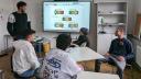 Fünf junge Erwachsene sitzen zusammen vor einem Smartboard. Darauf sind Abbildungen des Nutri-Scores zu sehen. Einer der jungen Erwachsenen steht neben der Tafel und spricht zu den anderen.