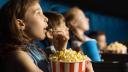 Ein Mädchen sitzt in einem Kinosessel, schaut in Richtung Leinwand und isst Popcorn. im Hintergrund sind zwei weitere Kinder zu sehen.