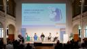 6 Personen sitzen in einem Saal auf einer Bühne, darüber eine Grafik mit dem Titel Leben lernen auf dem Lehrplan und der Unterschrift Wie steht es um Verbraucherbildung an Deutschlands Schulen